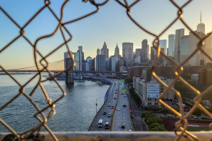 The lower Manhattan skyline with the sun setting behind it as seen through a hole in chain link fencing on the Manhattan Bridge